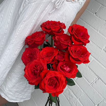 Bouquet of red roses held by a person wearing a white top against a white brick wall.