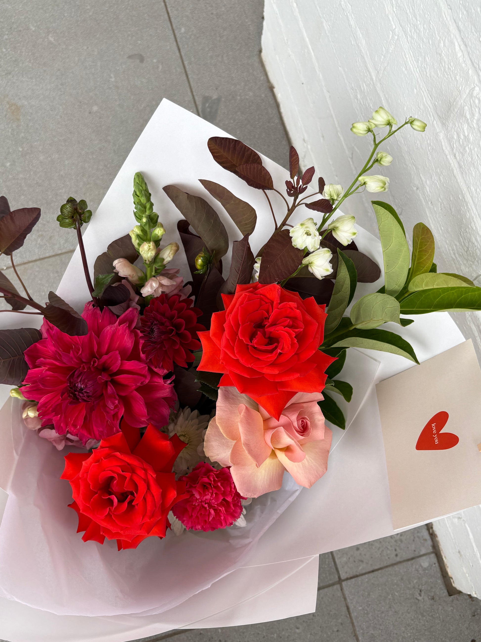 Bouquet of red and pink flowers with a card featuring a heart on a gray floor.