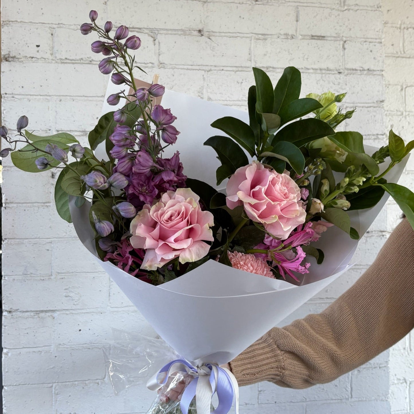 Bouquet of pink and purple flowers held by a person against a white brick wall.