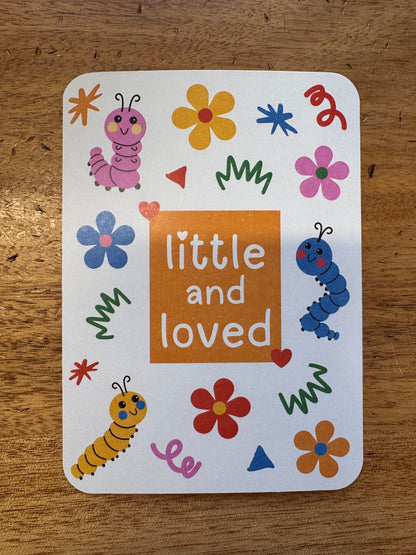 A square white card with rounded corners featuring the phrase 'little and loved' in white text inside an orange center block, surrounded by colorful flowers, caterpillars, and doodles, placed on a wooden surface.