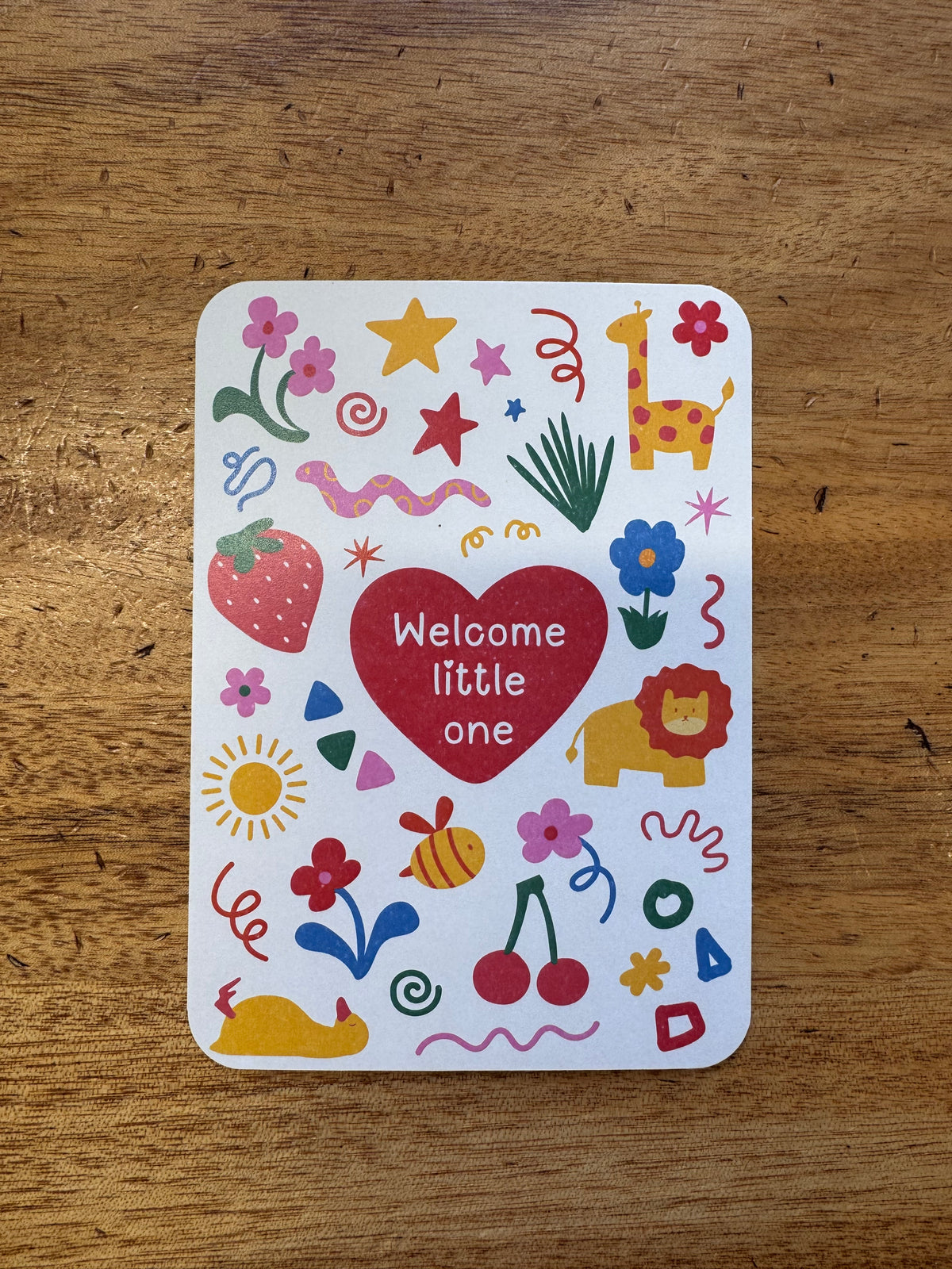 A square white card with rounded corners featuring a colorful doodle-style design and the words 'Welcome little one' inside a red heart, placed on a wooden tabletop.