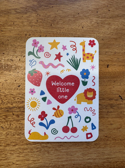 A square white card with rounded corners featuring a colorful doodle-style design and the words 'Welcome little one' inside a red heart, placed on a wooden tabletop.