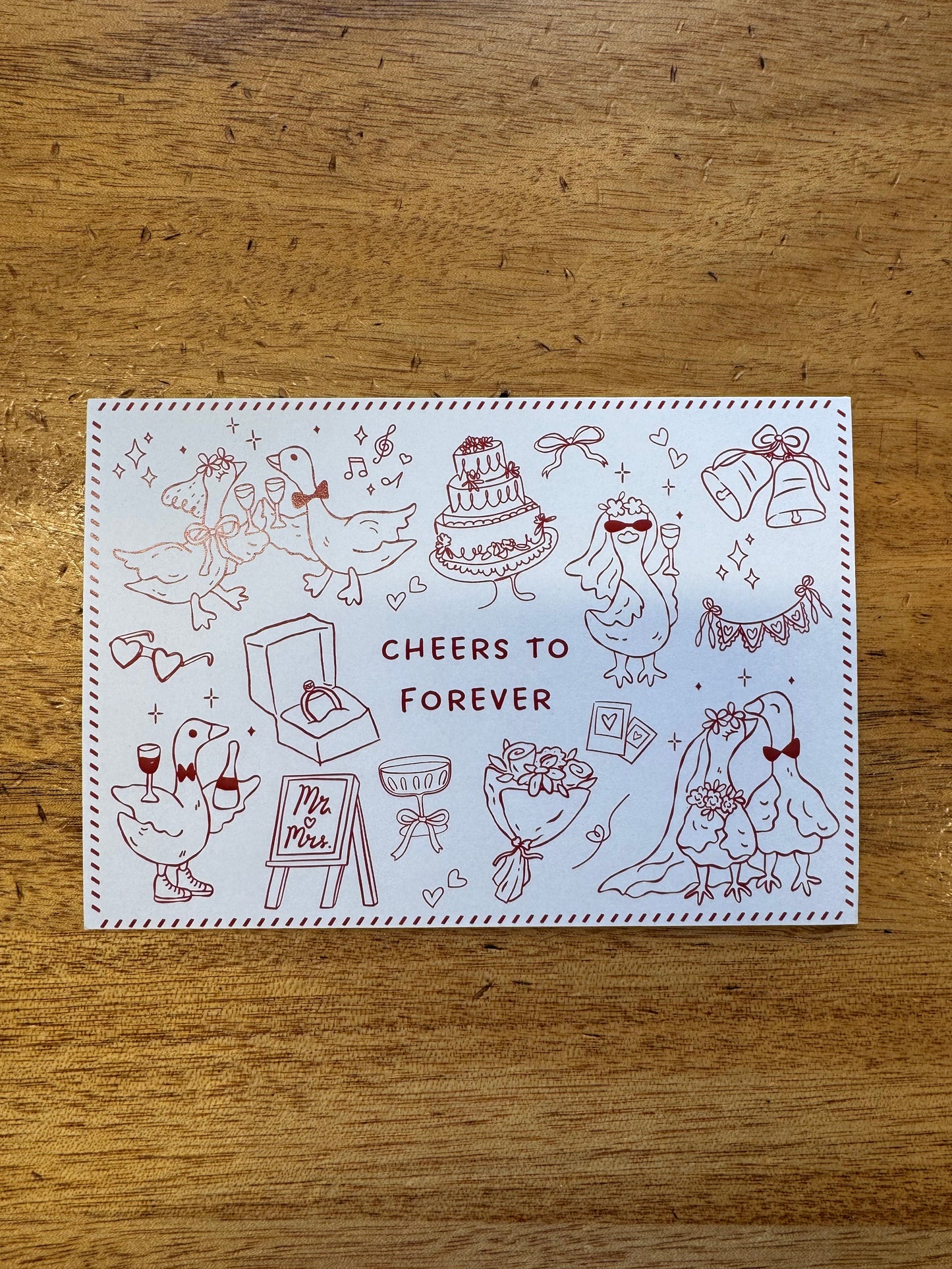 A square white greeting card with red line-art illustrations of ducks and the words 'CHEERS TO FOREVER' in the center, photographed on a wooden tabletop.