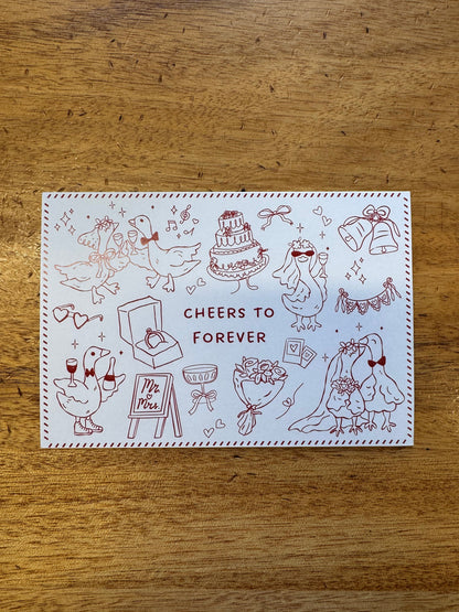 A square white greeting card with red line-art illustrations of ducks and the words 'CHEERS TO FOREVER' in the center, photographed on a wooden tabletop.