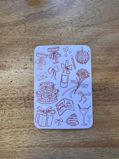 A rectangular white card with rounded corners featuring brown line-art illustrations of party and celebration motifs, placed on a wooden surface.